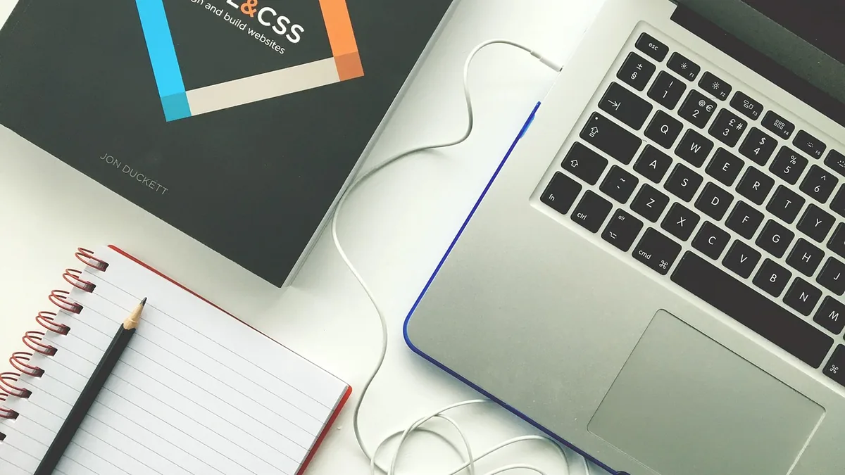 Dizüstü bilgisayar, HTML kitabı, kulaklık ve not defteri bulunan masa